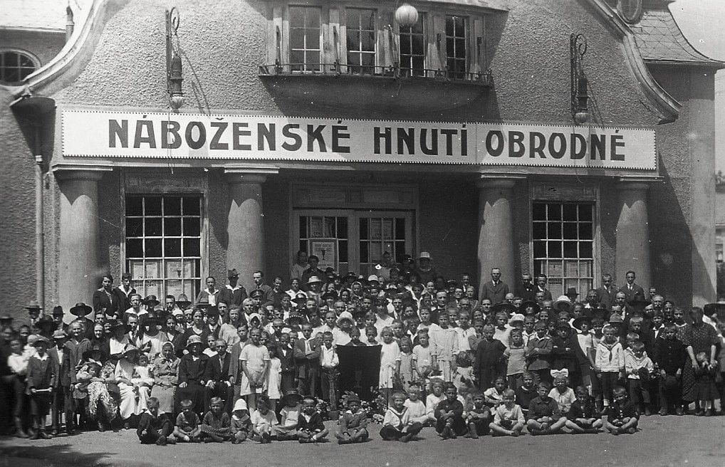 Skupinová fotografie více než stovky lidí všeho věku před BIO Elite Jihlava. Nad vchodem do budovy je velký nápis NÁBOŽENSKÉ HNUTÍ OBRODNÉ. Černobílá fotografie je z roku 1923.